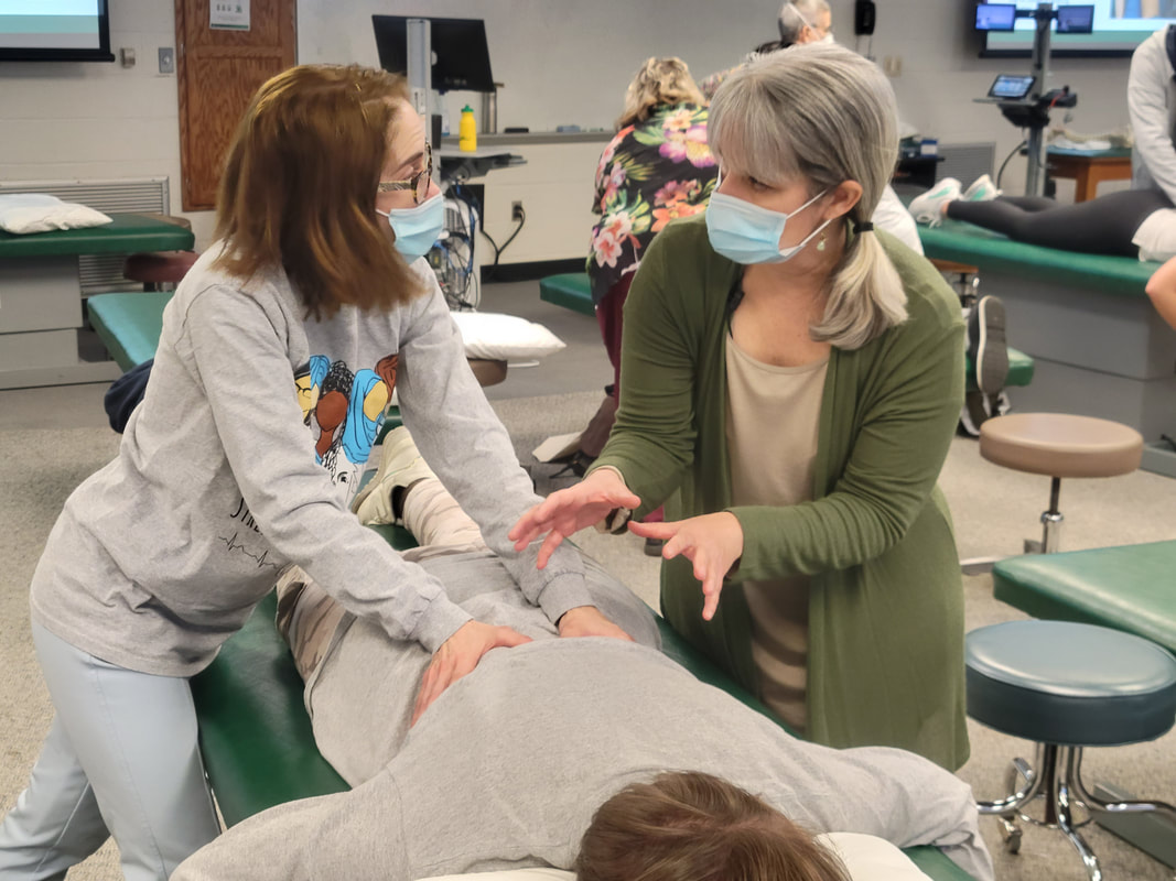 Instructor teaching resident a manual medicine technique with resident hands on middle back. Both are standing on either side of patient table while patient is face down.